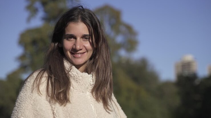 Retrato 4K de una joven y encantadora chica mirando a cámara y sonriendo, iluminada por el sol, con cielo azul y árboles de fondo, primer plano