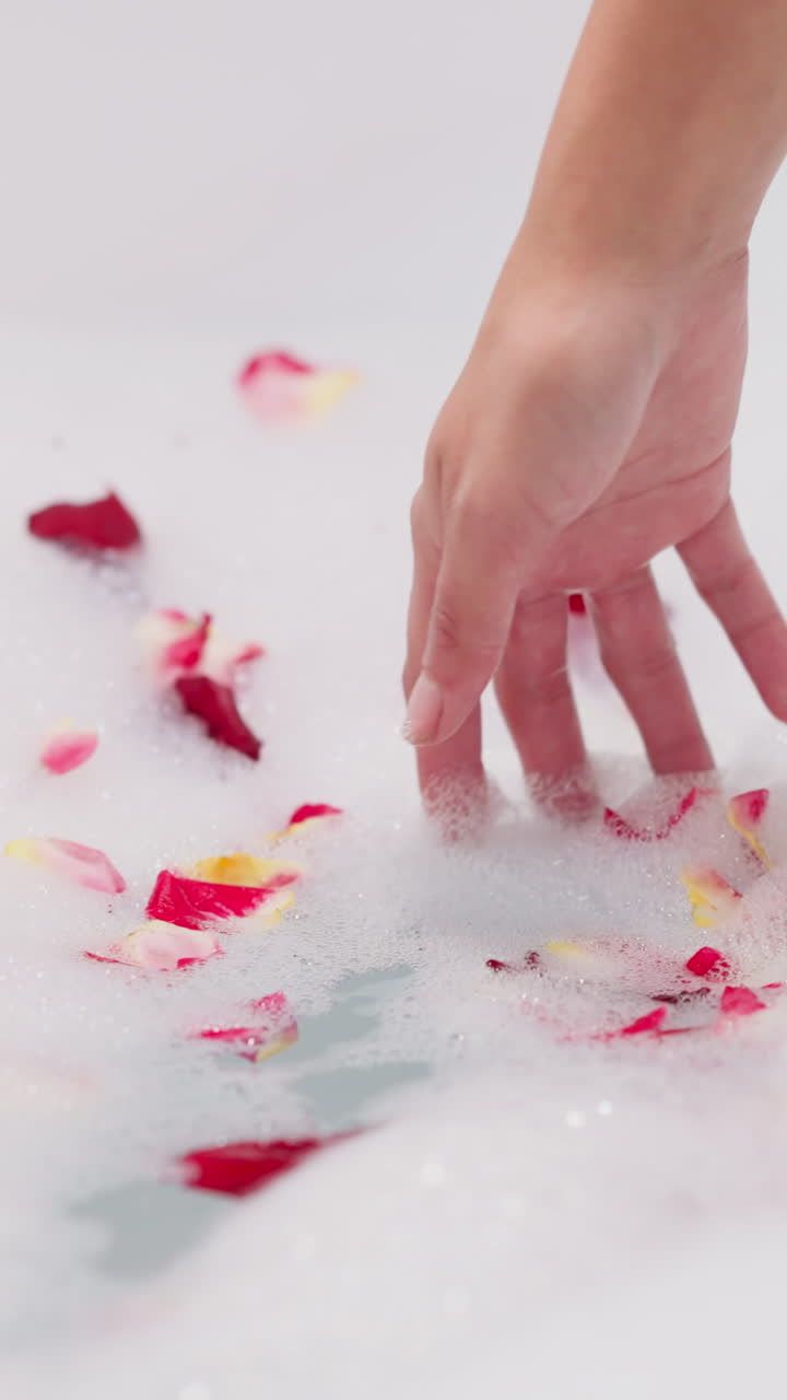 Lady dips hand into foam with rose petals closeup. Woman prepares romantic bath with fresh flowers stirring foam in bathtub. Idea for Valentine day
