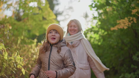 mother following daughter in forest path while child runs joyfully under golden sunlight glow around head, foliage framing pathway, autumn leaves shimmering as family enjoys playful outdoor moment