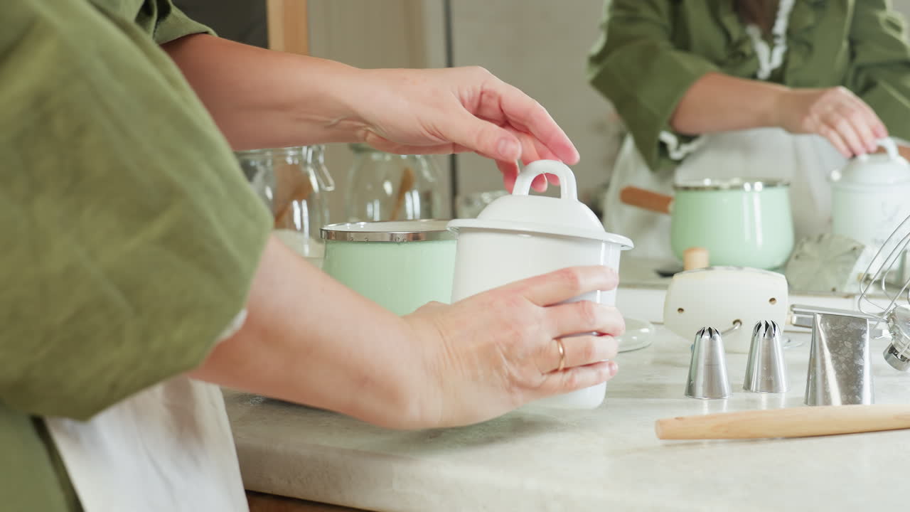 School girl in green dress and apron places white pot on kitchen scale, opens it, and drops lid beside it while preparing to lift pot, with mirror reflecting actions