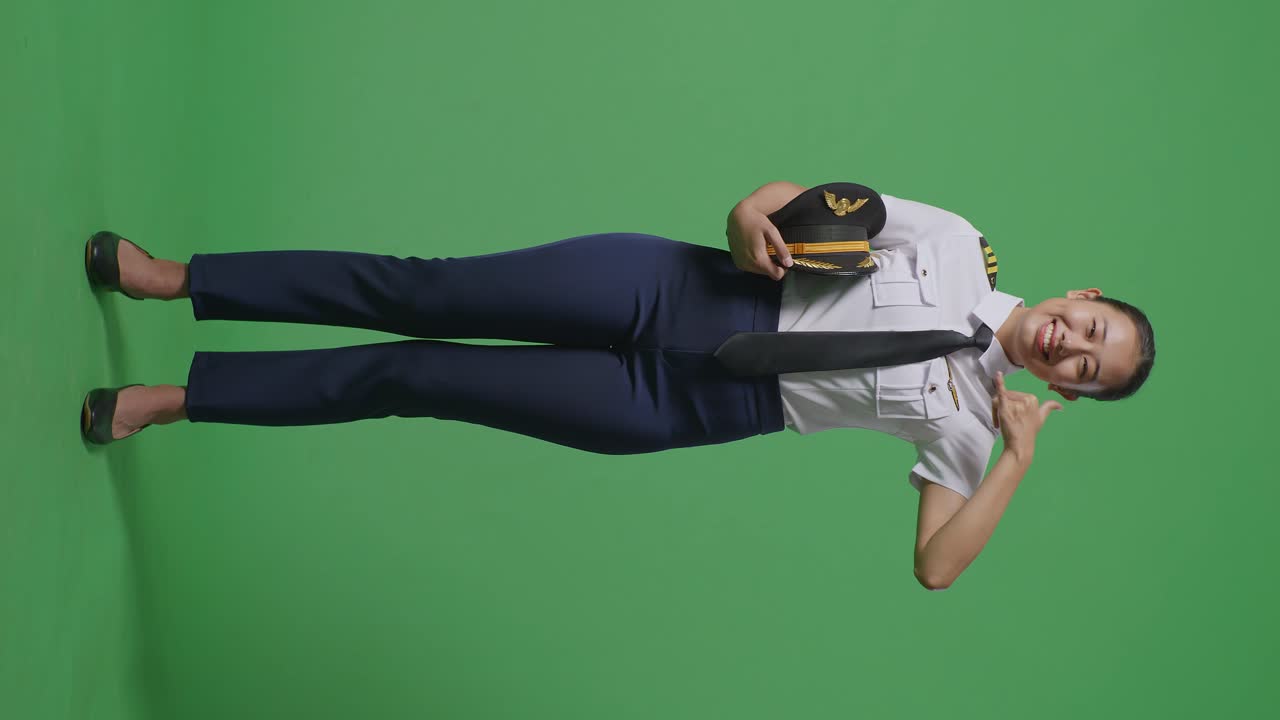 아시아 여성 조종사가 미소 짓고 녹색 스크린 배경 스튜디오에 서있는 동안 카메라에 제스처를 호출합니다.