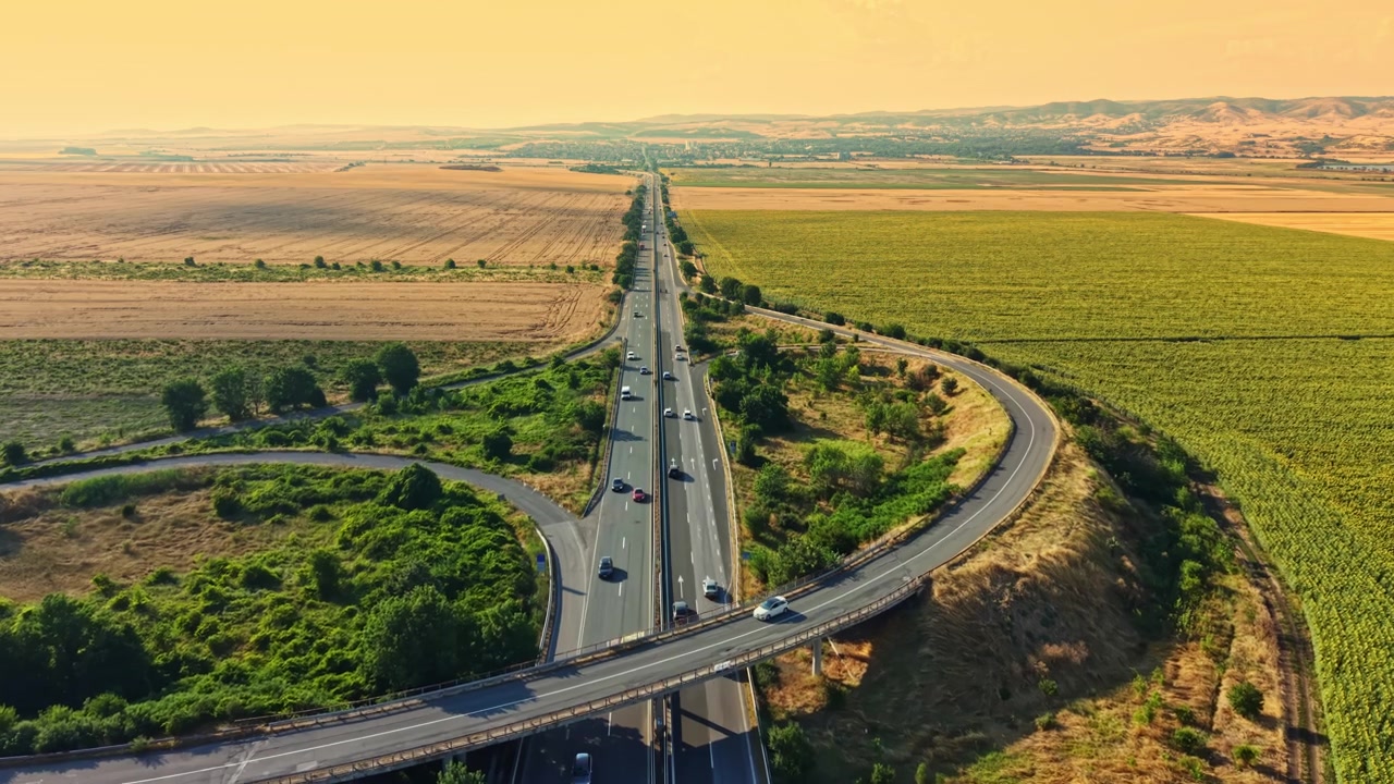 Un'ampia strada attraversa campi in una zona rurale con colline in lontananza in una giornata di sole.