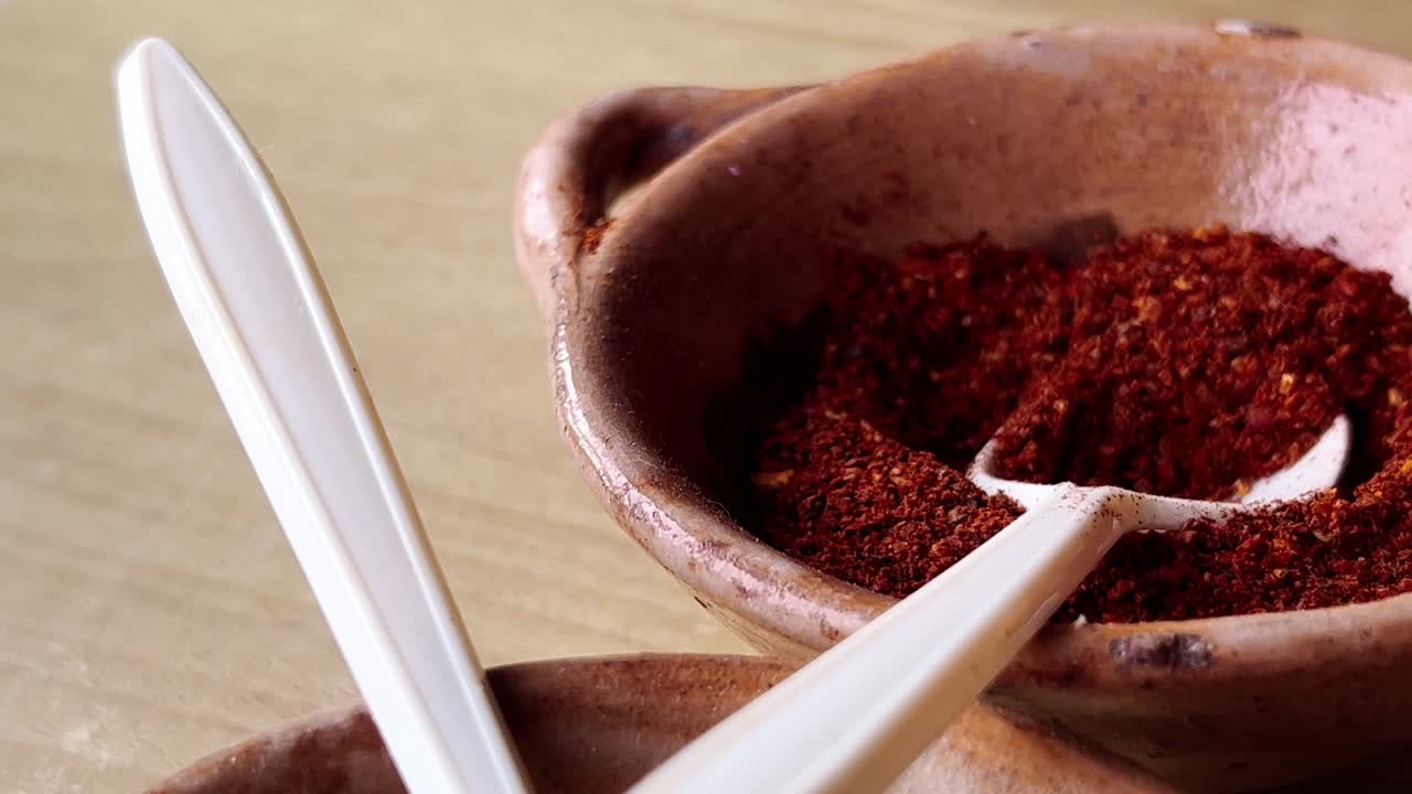 Plastic spoons scooping oregano and chili powder from rustic brown clay bowls, showcasing the vibrant colors and textures of traditional Mexican pozole preparation
