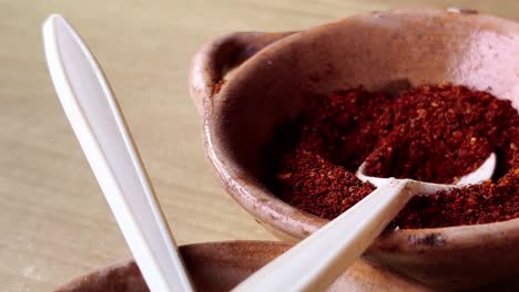 plastic spoons scooping oregano and chili powder from rustic brown clay bowls, showcasing the vibrant colors and textures of traditional mexican pozole preparation