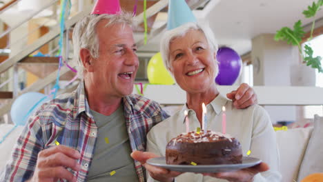 feliz pareja de adultos mayores caucásicos en sombreros de fiesta haciendo video llamada de cumpleaños con pastel y confeti de oro