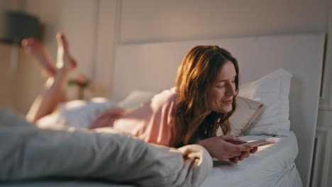 young woman using mobile phone at home. smiling happy female lying in bed typing