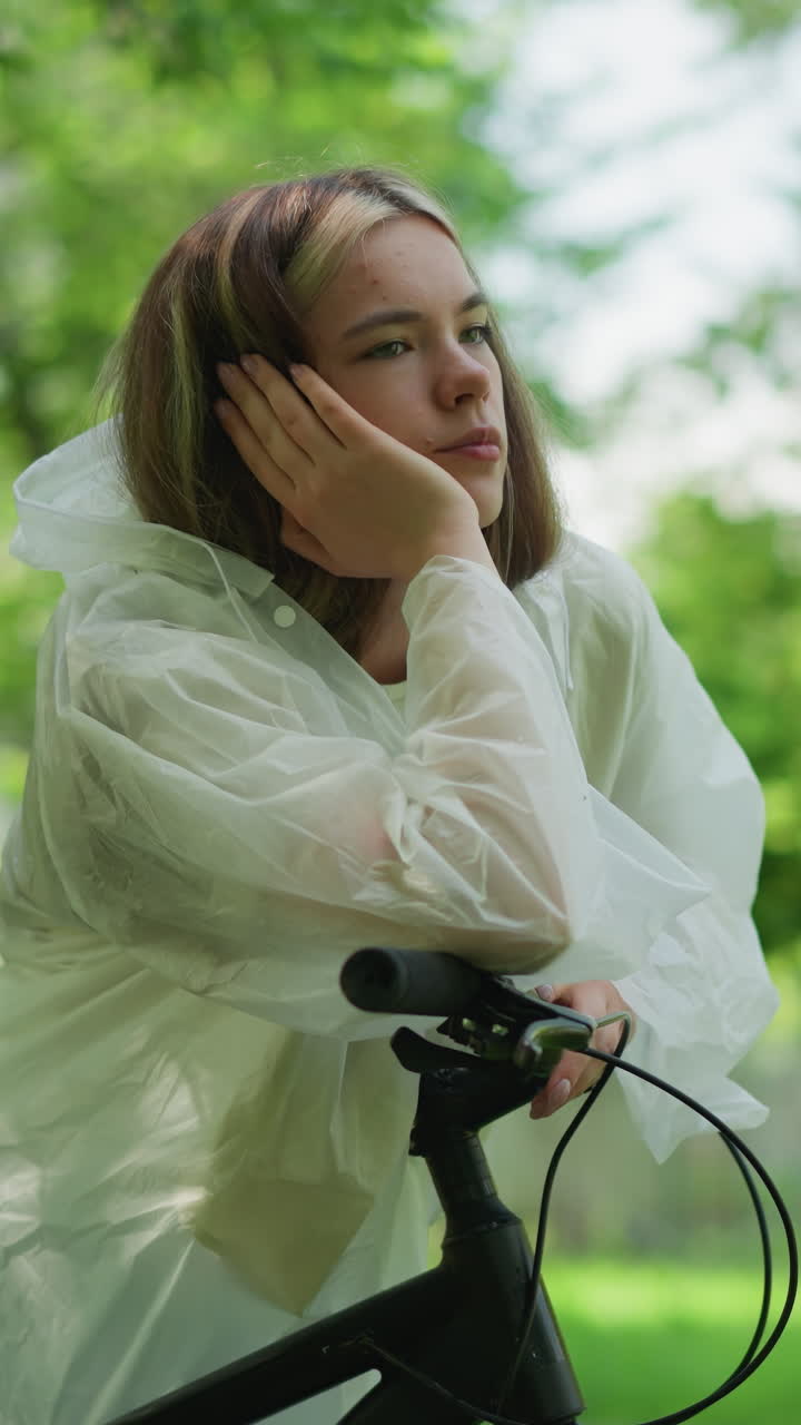 반투명한 레인코트를 입은 젊은 여성은 그녀의 손에 머리를 고, 그녀의 자전거 손잡이에 사려 깊게 기대고, 상에 빠져, 배경에는 울창한 녹색 나무가 있습니다.