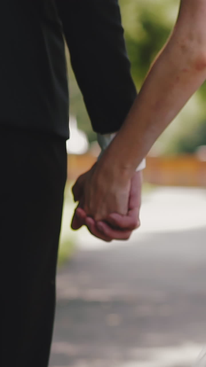 CU shot, slow motion: elegant young woman in tight lacy wedding dress and man in suit walk joining hands along summer park extreme close backside view