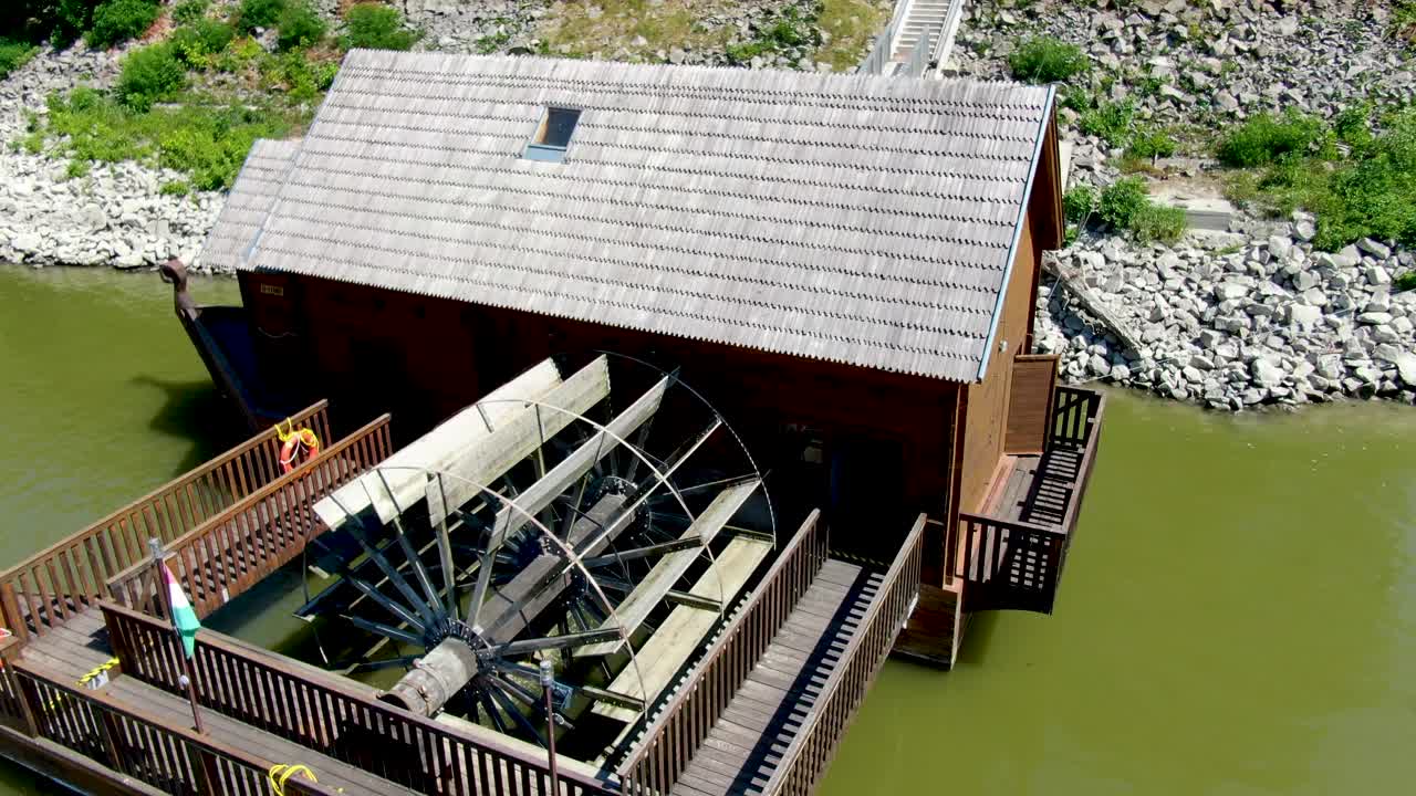 Tracking aerial past large water wheel on ship mill, a floating lumber mill powered by Danube River.