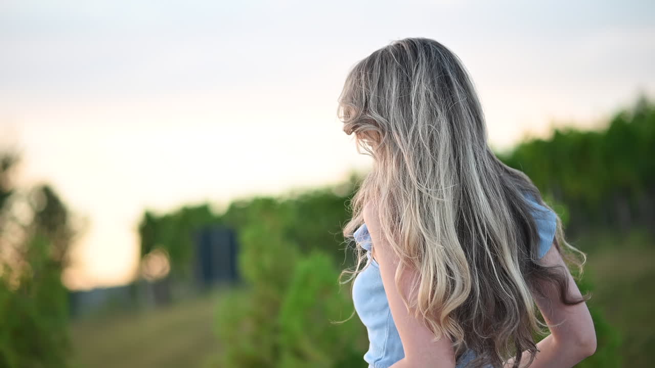 Young woman standing outdoors with her hair blowing in the wind, smiling peacefully at sunset