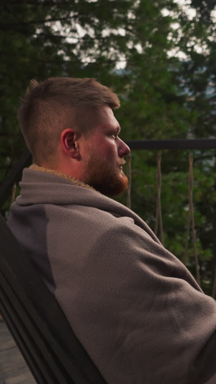 Man enjoys nature view from glamping terrace. Bearded guy wrapped with plaid sits in comfortable armchairs by round tent house. Relaxation in nature