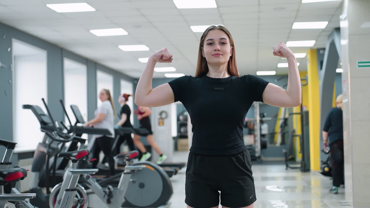 young woman flexes arms to show muscle definition and confidence in modern gym with people on treadmills and exercise machines, highlighting energetic active lifestyle