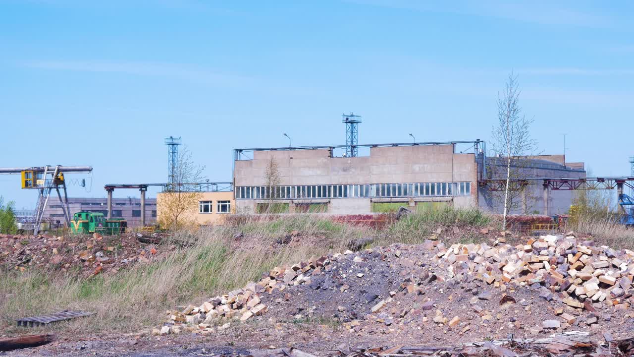 вид со стороны на заброшенный советский завод по выплавке продукции тяжелой металлургии на территории лиепаяс металлургс, груды мусора и хлама на переднем плане, дальние кирпичные здания, солнечный день, крупный план