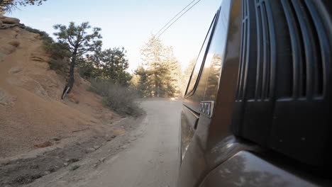 Close-up-view-of-the-side-of-a-black-car-while-on-the-way-to-San-Bernadino-NF,-Big-Bear-Lake,-CA,-USA-with-trees-at-sunset