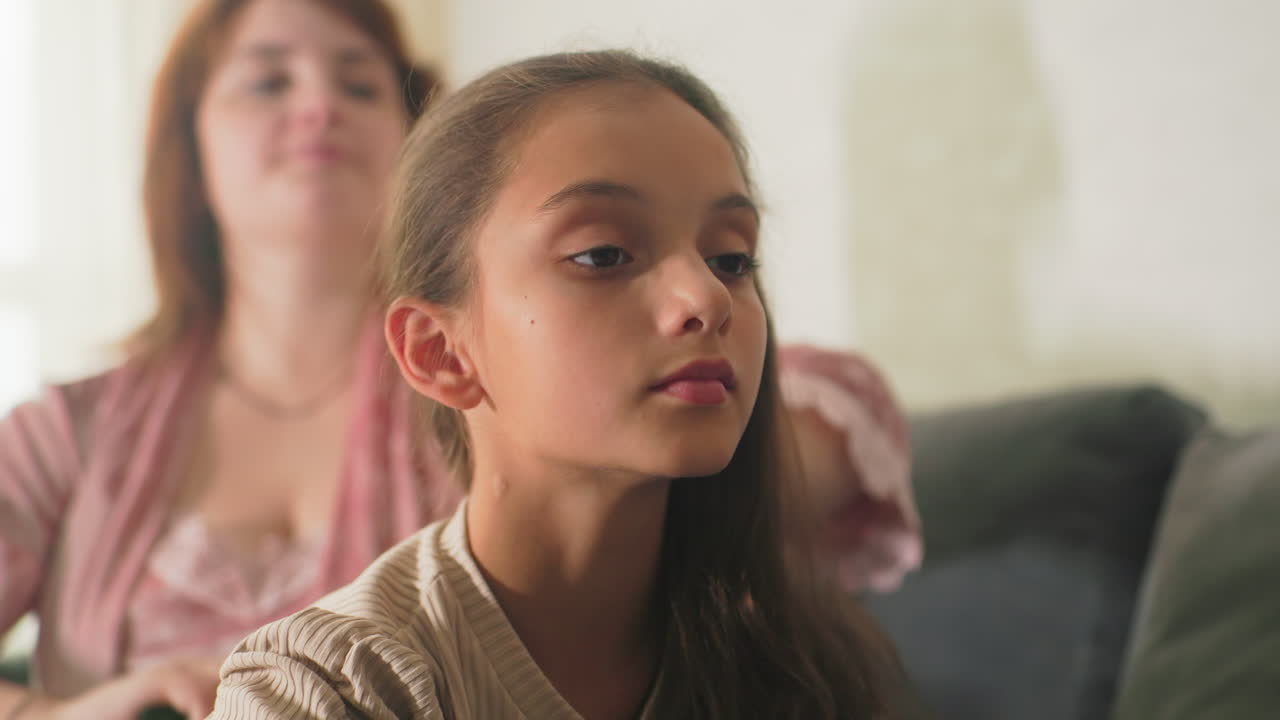 Mother brushing daughter’s hair on couch in cozy living room. Peaceful, intimate moment between parent and child, filled with affection, calm, and soft light coming through window