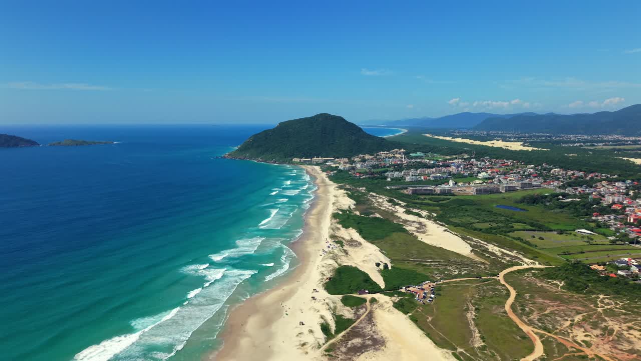 Rastreamento aéreo em alta altitude sobre a Praia do Santinho. O clipe mostra ondas quebrando na areia branca com uma área residencial próxima e o Oceano Atlântico sob céus tropicais límpidos