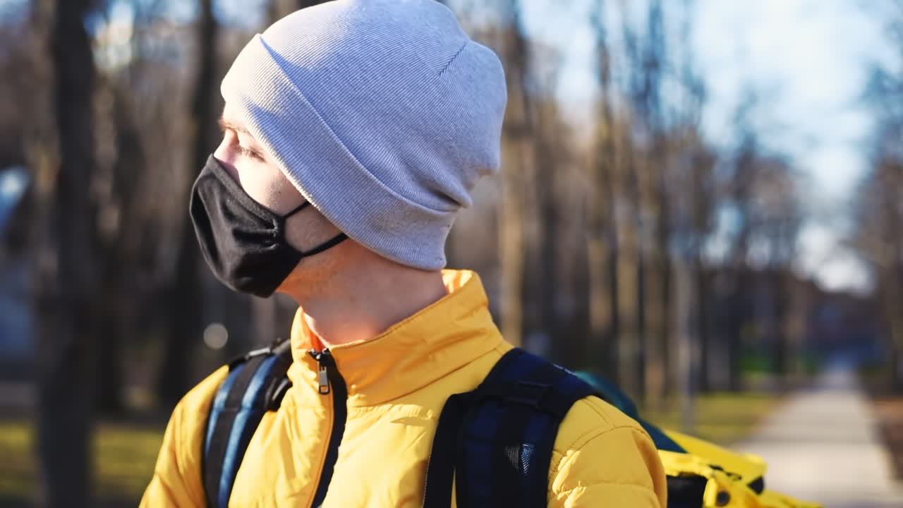 Food delivery man in a park looking into camera. Black medical mask, yellow backpack and jacket. Winter