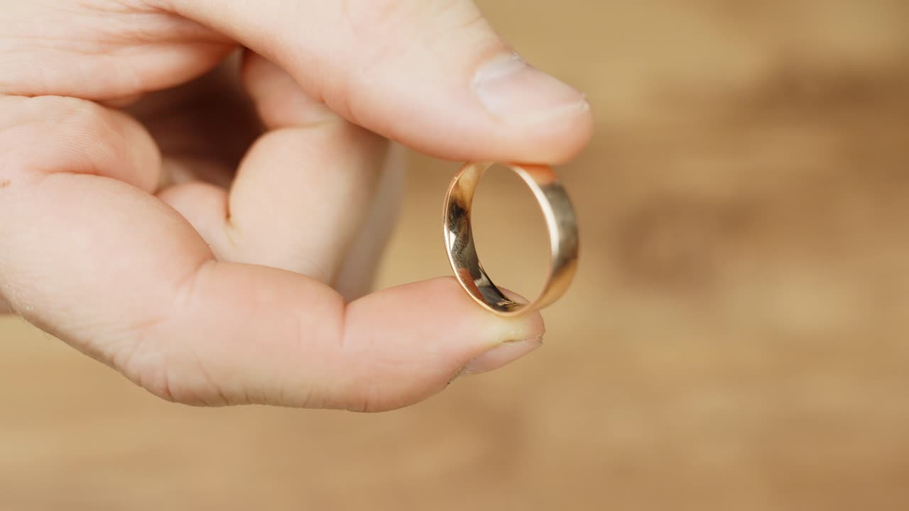 Close up person's holding a plain, polished gold wedding ring against wooden table in blurred background