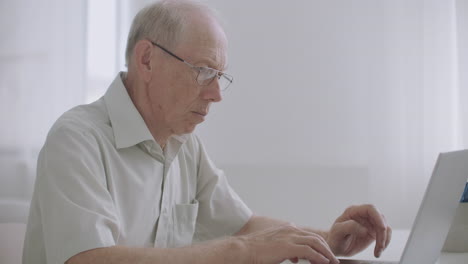 Un-Hombre-Gris-De-Edad-Avanzada-Con-Gafas-Está-Trabajando-Con-Un-Portátil-Navegando-Por-Internet-Y-Chateando-En-Línea-Escribiendo-Mensajes-En-El-Teclado