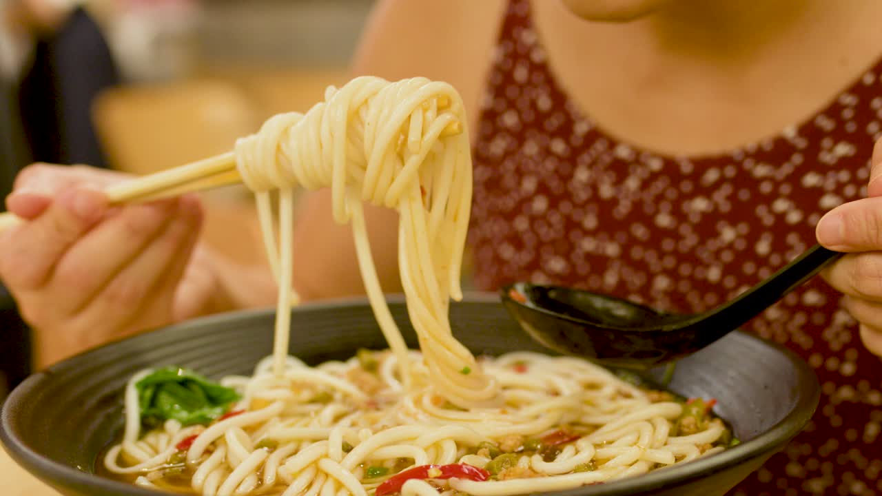 Close-up of woman eating spicy noodle soup with chopsticks in warmly lit restaurant setting
