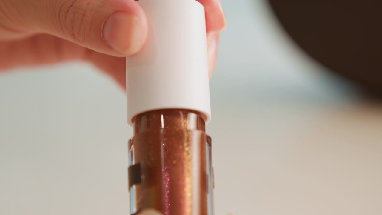 Close-up motion of doe-foot wand rising from clear bottle filled with shimmery brown-gold cosmetic liquid, with viscous product stretching from tip, glossy residue on wall, soft studio light