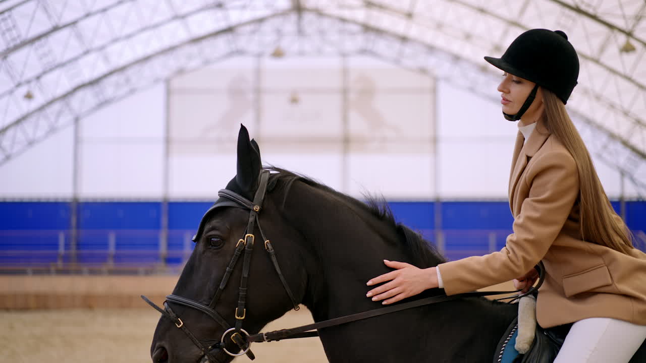 Female jockey in black helmet and beige jacket riding a beautiful black horse. Woman calms down the animal caressing it. Blurred backdrop.