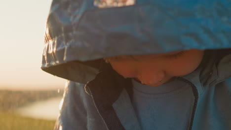 un niño con un impermeable mira hacia abajo con una expresión triste mientras está de pie bajo la lluvia.