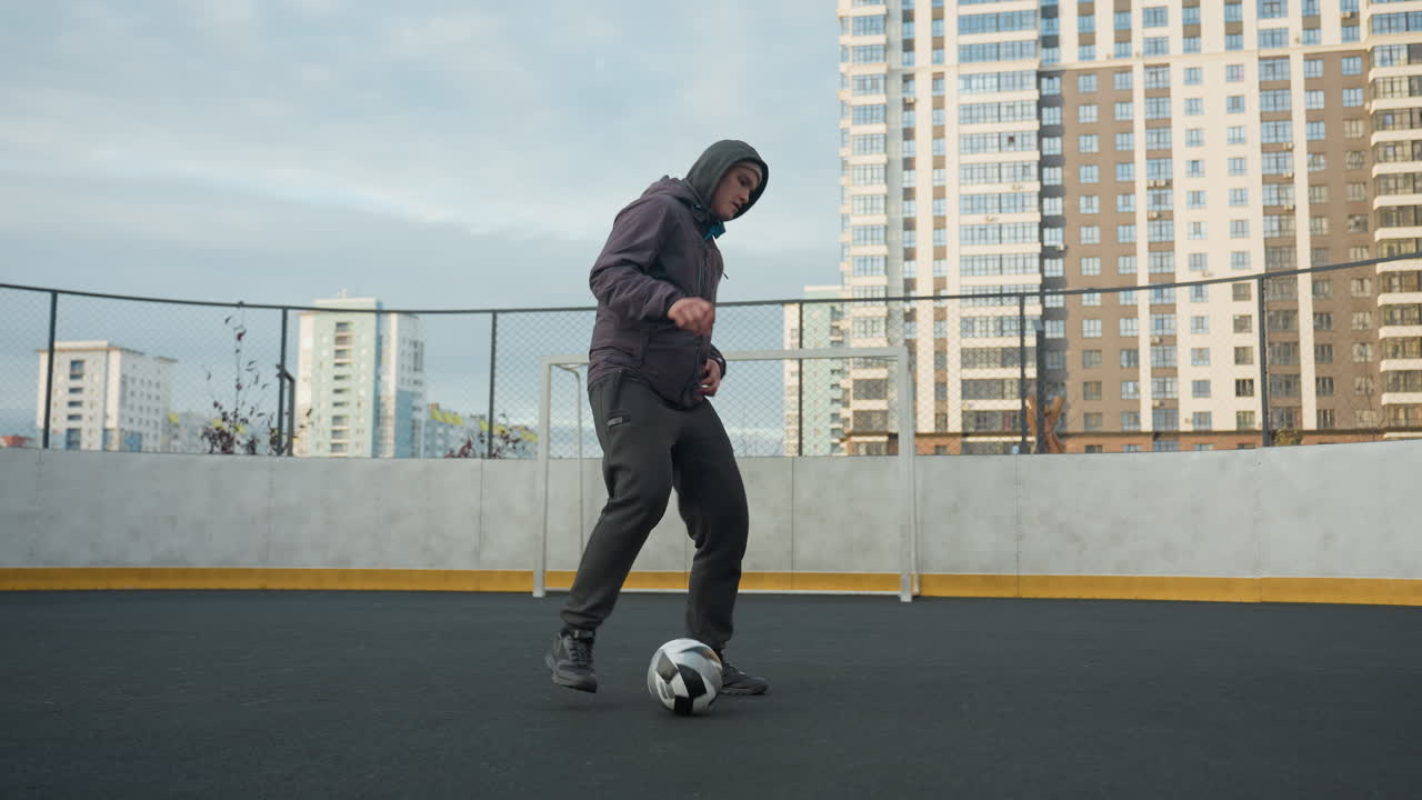 訓練中にスポーツアリーナで足でサッカーボールを巧みに制御している男性背景は都市の建物ゴールポストフェンシングを展示しています