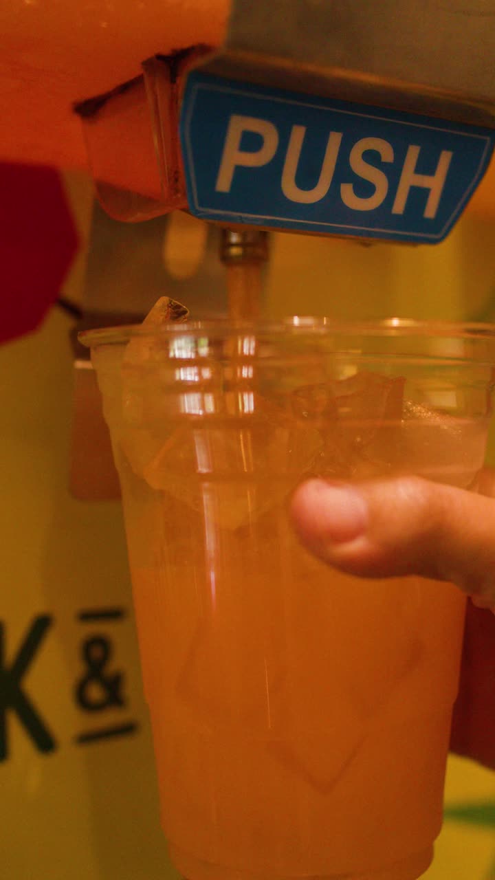 Una mano preme un pulsante per versare una bevanda all'arancia con ghiaccio in un bicchiere di plastica da un distributore automatico.
