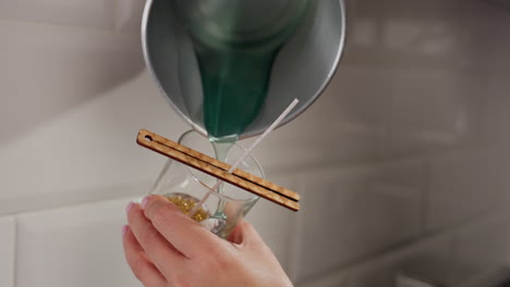close-up of individual pouring blue liquid into glass cup containing golden candle wax and centered wick held by wooden sticks, capturing precise moment of craft process in modern indoor kitchen