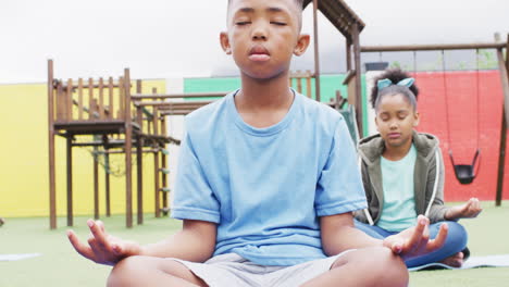 Diverse-schoolchildren-exercising-and-meditating-on-mats-at-school-playground