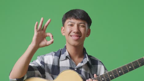 close up of asian teenager with guitar showing okay gesture and smiling on green screen background