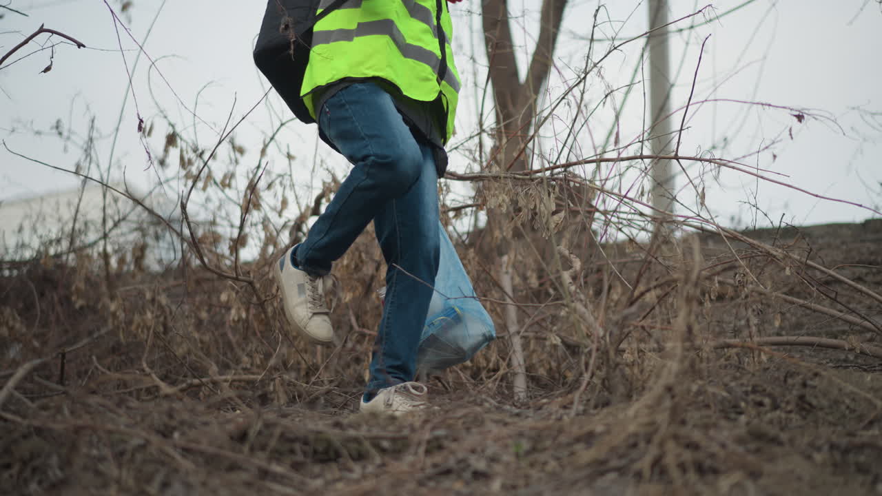 Volunteer wearing reflective safety vest and hoodie collecting litter with gloved hands into blue plastic bag among dry branches and leaves during environmental cleanup in natural outdoor area