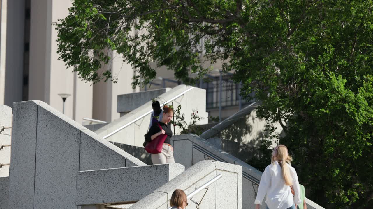 Una scala in cemento del campus è percorsa da pendolari mentre una singola persona scende verso una piazza sotto un albero.