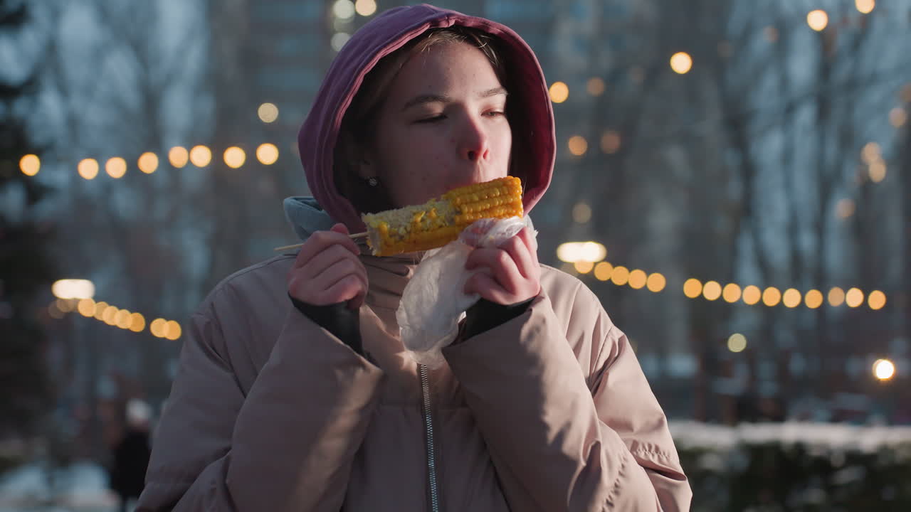 색 봉투에 싸여있는 막대기에 옥수수를 즐기는 젊은 여인, 흐릿한 배경에서 보케 조명, 신선한 옥수수에서 증기가 상승, 겨울 야외 간식 시간 동안 배경에서 는 사람들