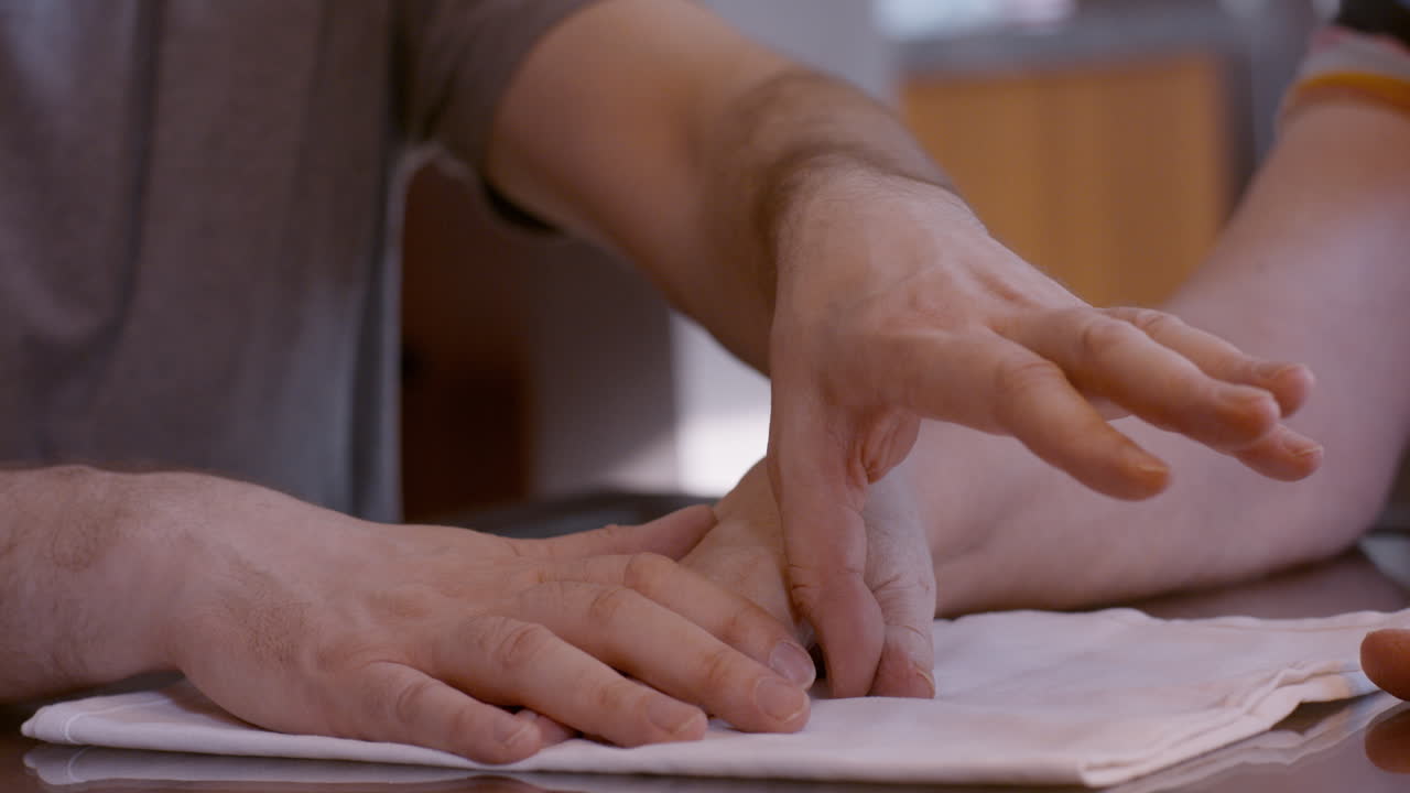 Middle-aged man caregiver assists senior woman stroke survivor by gently stimulating and mobilizing her hemiplegic right thumb during a hand therapy session at home kitchen table, on cloth pad.