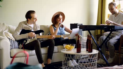 charming female vocalist is singing looking at paper with lyrics while her handsome male friends are playing keyboard and guitar. table with bottles and snacks is visible.