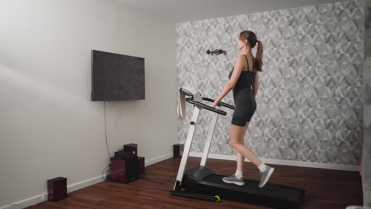 Side view of confident lady walking majestically on treadmill at home, maintaining composed posture while focused on workout, dressed in dark athletic wear, towel draped over handle