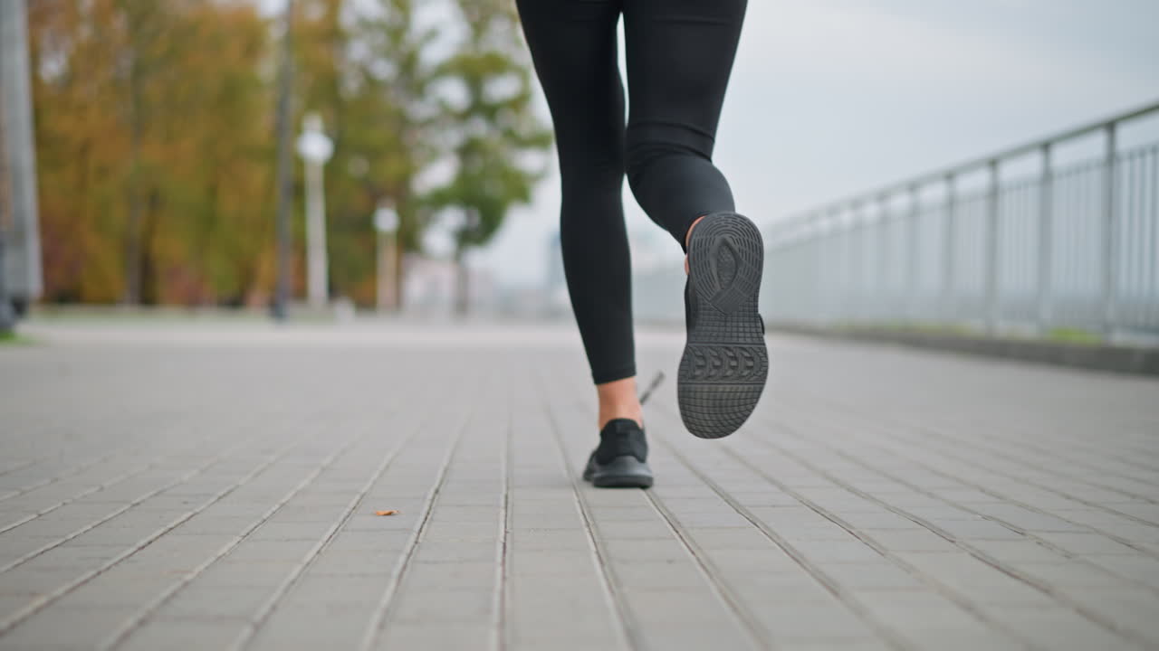 Close-up view of woman jogging in sneakers along paved street path near iron fence with blurred background showing trees and distant landscape under cloudy sky