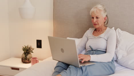 senior woman using laptop in bed, focusing on work at home