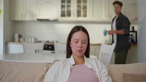 Brunette-pregnant-girl-sits-on-the-sofa-while-her-husband-brings-her-a-cup-of-hot-tea-at-home-during-the-weekend