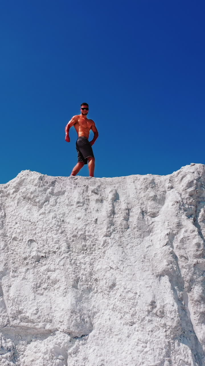 Drone view on the high white mountain and muscular man on top of it in summer. Topless young sportsman showing his trained body while standing on the top of the hill under blue sky. Vertical video