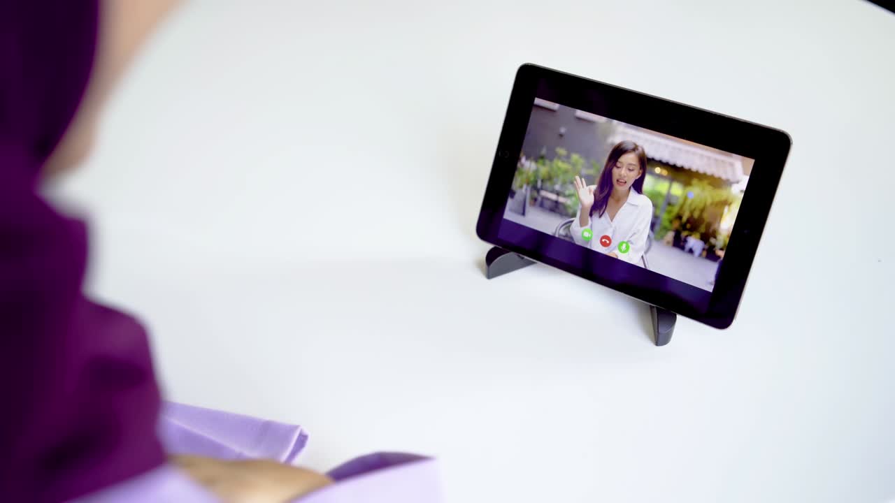 vista trasera de una joven musulmana sentada en una mesa en la habitación negociando usando una tableta y una aplicación de video llamada, agitando la mano haciendo un gesto de saludo, teniendo una reunión en línea de video llamada con una amiga