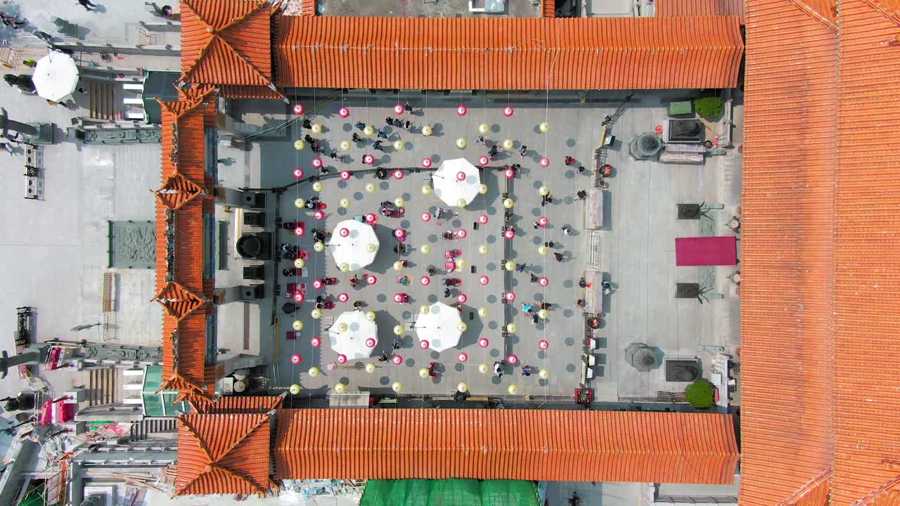 Wong Tai Sin temple court with people lighting incense sticks and kneel before the main altar under Chinese lanterns, Top down one minute aerial view.