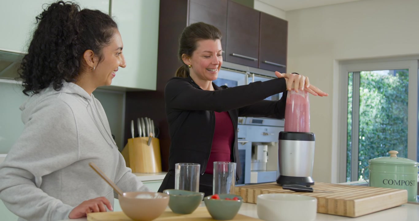 Diverse donne che osservano in cucina mentre la donna a destra preme il bicchiere del frullatore preparando un frullato rosa