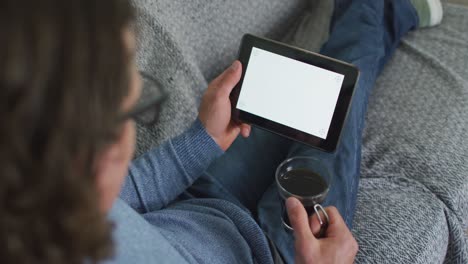 タブレットとコーヒーを飲みながらリビングのソファーに座っている白人男性