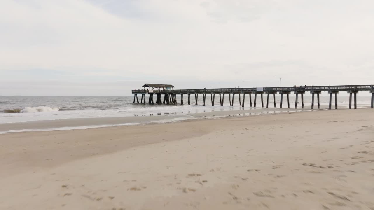 Drone di una spiaggia vuota a Tybee Island che si avvicina al molo e all'oceano con onde agitate, a bassa quota sulla sabbia