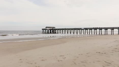 drone of an empty beach in tybee island approaching pier and ocean with rough waves, low to the sand