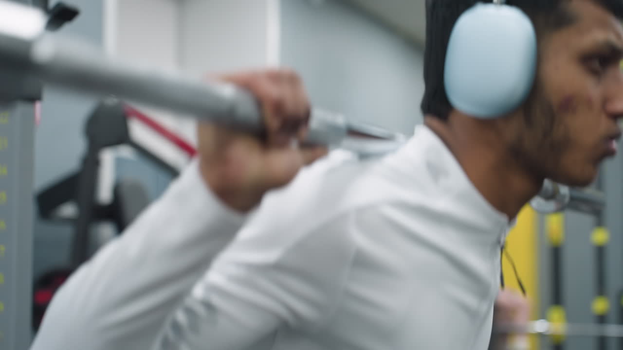student carrying barbell across shoulders while performing intense squat workout under pressure in indoor gym environment with onlookers in background and bright modern fitness equipment visible