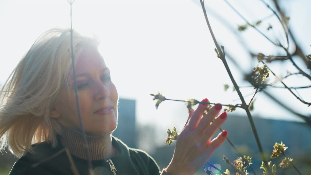 Blonde woman with short hair wearing dark jacket and turtleneck touching budding branches with care on sunny spring day outdoors, observing nature with gentle expression of seasonal growth